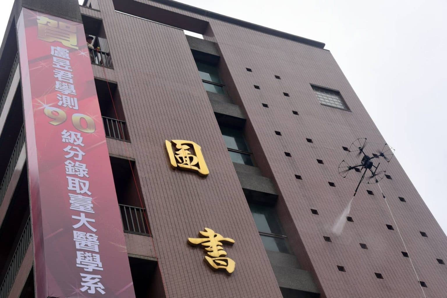 一般無人機創辦人江天農於僑泰高中圖書館大樓演示無人機外牆清洗技術 - 多旋翼無人機正在對棕色瓷磚外牆進行高空清潔作業，建築物上有「國書」金色大字